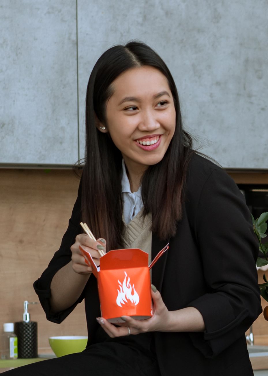 Smiling Asian woman using chopsticks to eat from a takeout box during lunch break indoors.